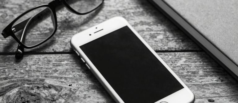 Black and white flat lay of a smartphone, eyeglasses, and notebook on a rustic wooden desk.