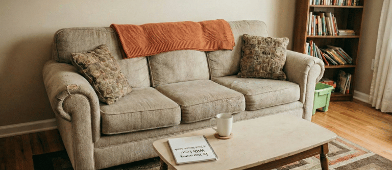 Cozy living room with a beige sofa, orange throw blanket, and a coffee table with a mug and book.