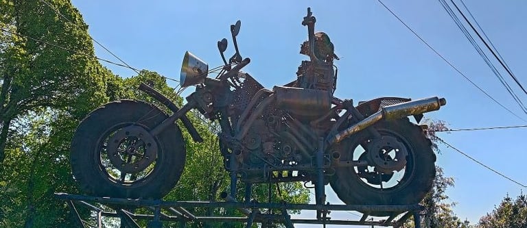A large rusty metal motorcycle sculpture made from scrap parts displayed outdoors against a blue sky.