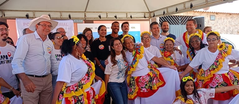 A group celebrates at a cultural event with women in traditional floral Baiana dresses and local community leaders.