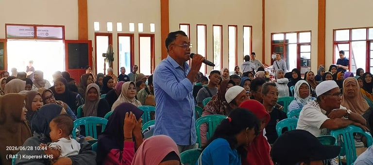 A man addresses a large group of people seated in rows, speaking into a microphone at a community event or meeting, with