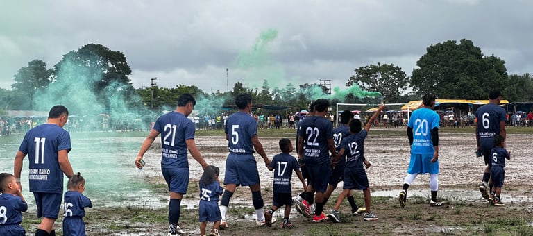 Soccer team and children walk across a muddy field with green smoke flares under a cloudy sky.