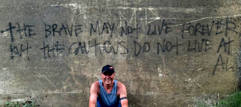 A man smiling in front of an inspirational graffiti quote on a concrete wall about bravery and living life.