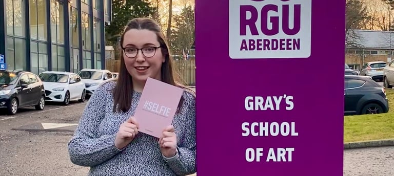 Erin, the owner of EB Socials, standing beside a sign for 'Gray's School of Art'