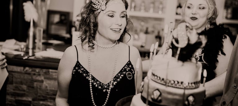 woman in 1920s outfit smiling at a birthday cake. another woman is smiling at the birthday girl.