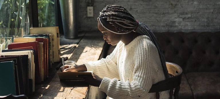 A young woman writes in a journal at a rustic wooden desk with books in a cafe.
