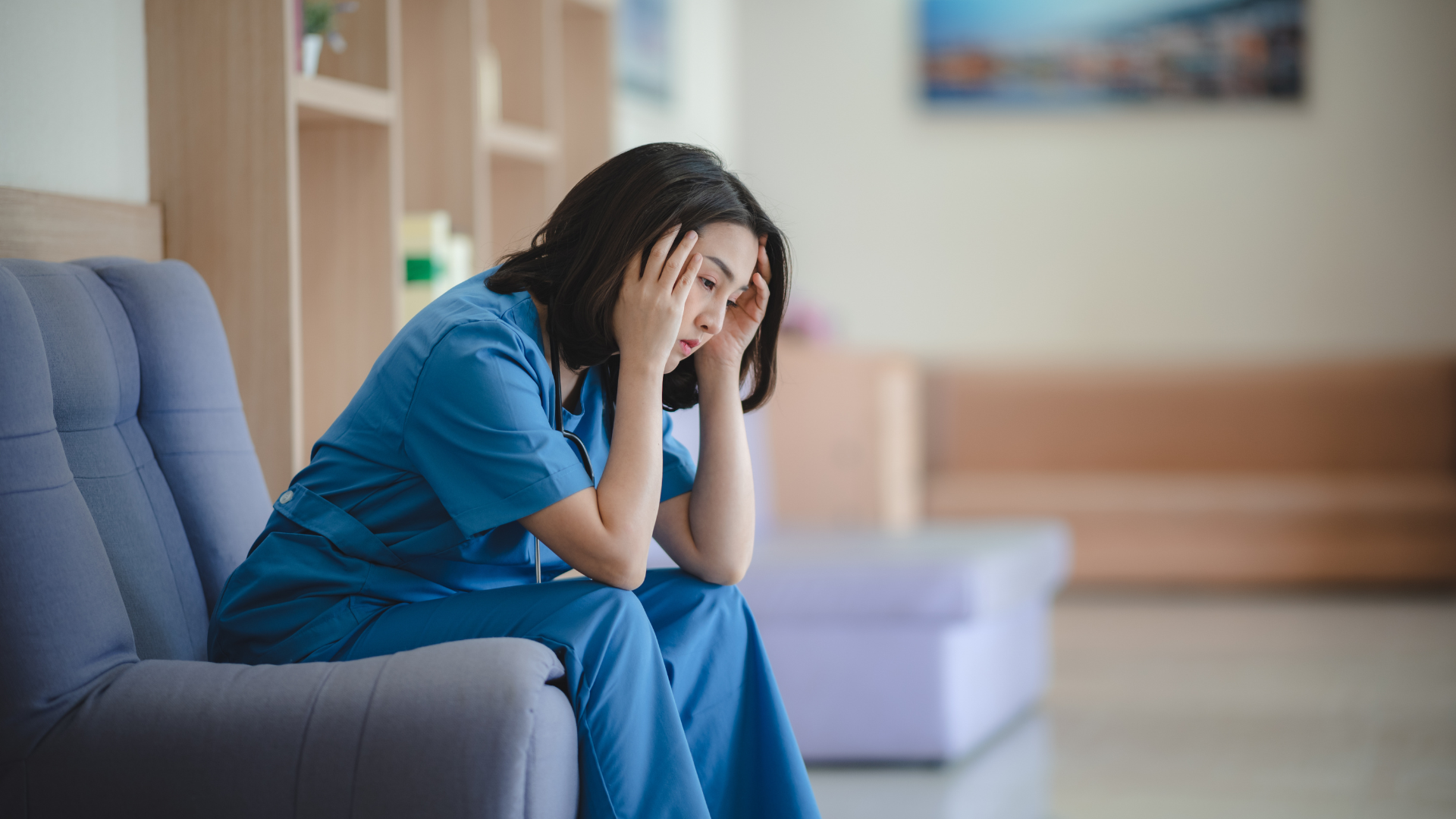 A stressed female nurse in blue scrubs sitting on a sofa and holding her head during a break.