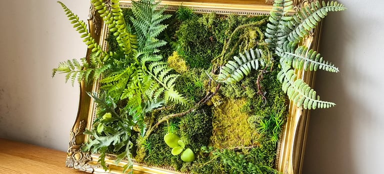 Framed preserved moss wall art with ferns in an ornate gold vintage frame on a wooden table.