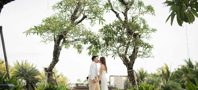 Couple standing under tropical trees during a romantic prewedding photoshoot at Apurva Kempinski Bali