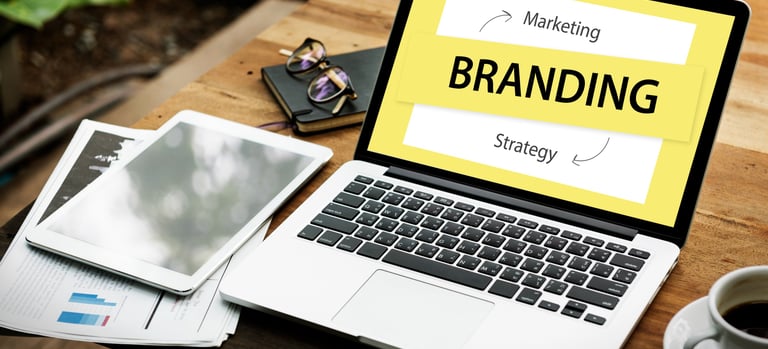 Notebook em uma mesa com tablet, café e cadernos, escrito "Branding Marketing Strategy" na tela. 
