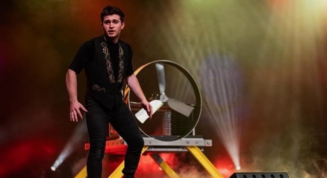 A male magician in a black vest performs on a stage with dramatic lighting and a large prop fan.