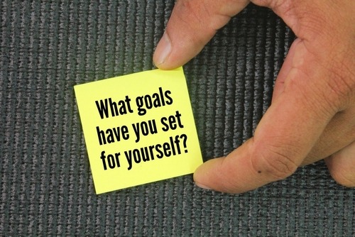Hand holding a yellow sticky note with goal setting motivation text on a grey textured background.