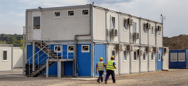 Two construction workers walk past a double-story modular office complex at a building site.