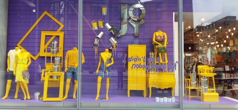 A vibrant purple and yellow retail window display featuring mannequins, paint buckets, and furniture.