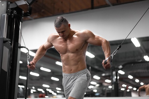 Muscular bodybuilder performing a standing cable chest fly exercise in a gym for pec development.