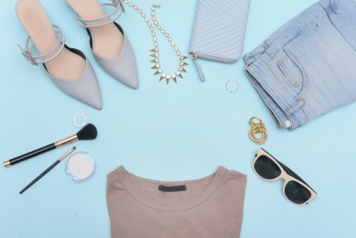 Flat lay of women's fashion accessories including heels, jeans, jewelry, and makeup on a blue background.