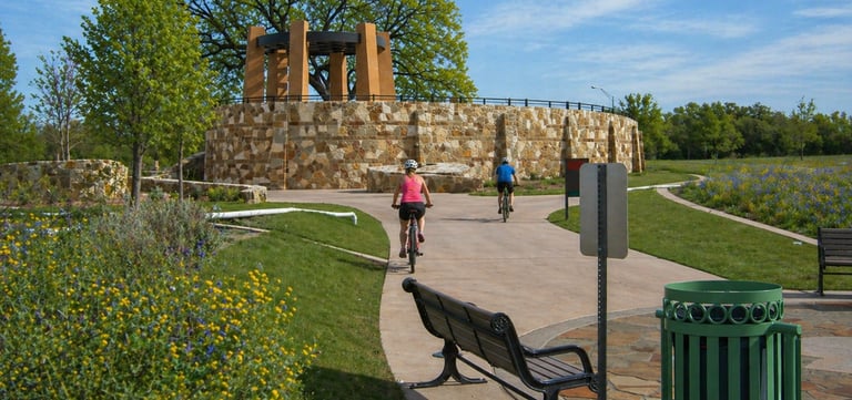 Cyclists ride on a paved trail near a stone pavilion on the San Antonio River Trail.