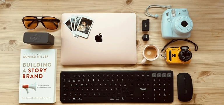 Flat lay of a workspace with a MacBook laptop, Instax camera, Trust keyboard, and Building a Storybrand book.