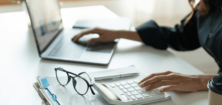 una mujer sentada en un escritorio con un portátil, una taza de café, una calculadora y sus gafas