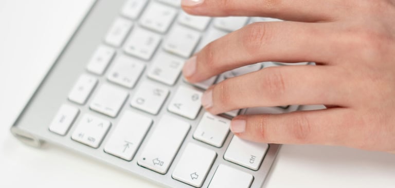 A person's hand typing on a slim silver and white wireless computer keyboard on a white desk.