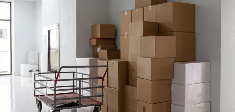 "Montón de cajas de cartón apiladas junto a un carrito de carga, listas para el traslado.