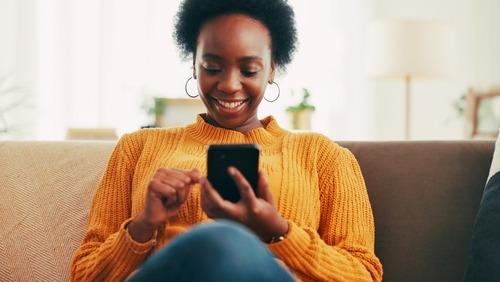 Smiling young Black woman in a yellow sweater using a smartphone while relaxing on a sofa.