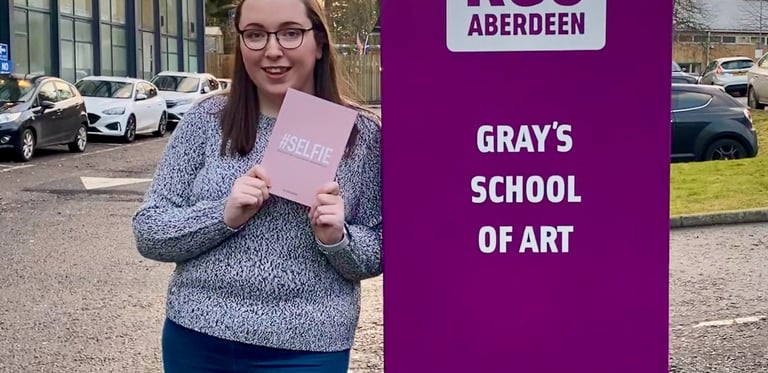 Erin, the owner of EB Socials, standing beside a sign for 'Gray's School of Art'