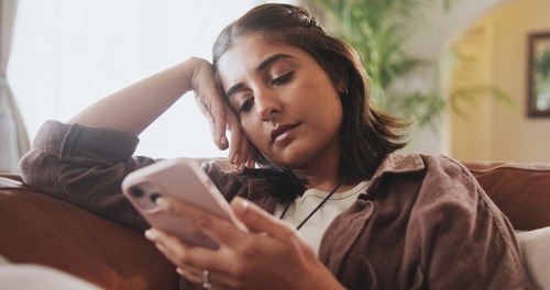 A bored woman lounging on a couch while scrolling through her smartphone at home.