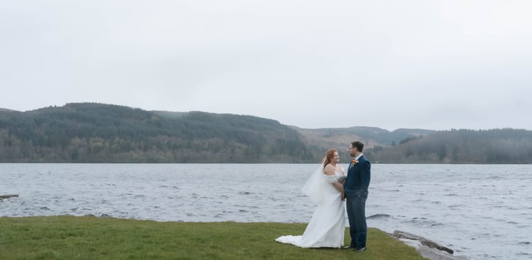 A bride and groom posing for wedding photos by a scenic lake in Scotland on a misty day.