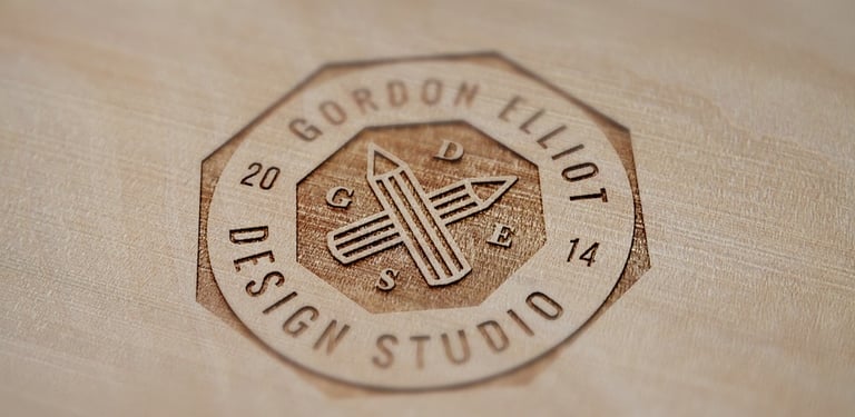 Laser engraved Gordon Elliot Design Studio logo on a light wood surface featuring crossed pencils.