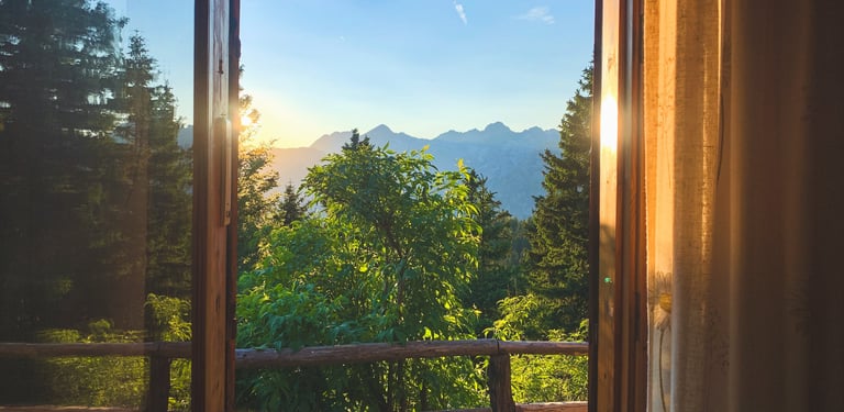 A scenic mountain sunset view through an open wooden cabin window surrounded by pine trees.