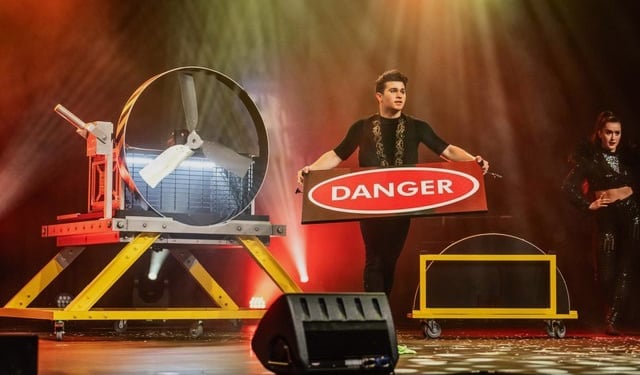 Magician Alfredo Lorenzo performing a dangerous stage illusion with a large industrial fan and a danger sign.