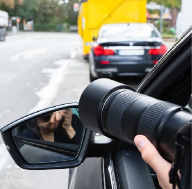 A private investigator uses a telephoto lens to photograph a car from a distance for surveillance.