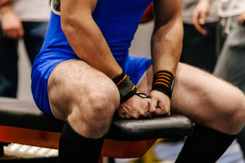 A powerlifter in a blue singlet wearing wrist wraps sits on a weight bench preparing for a lift.