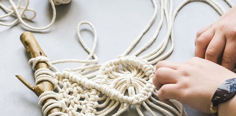 Manos creando un tapiz de macramé hecho a mano utilizando cuerda de algodón color crema y una rama.