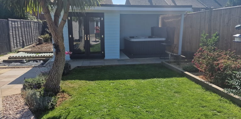 Modern garden cabin with a luxury hot tub and glass doors next to a green lawn and palm tree.