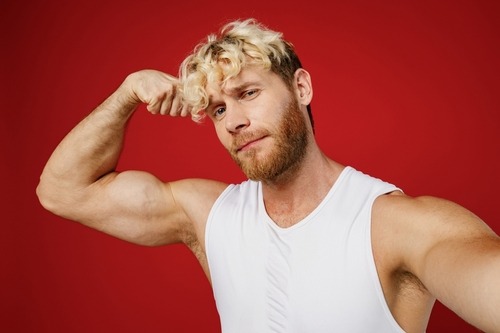 A blonde man with a beard flexes his bicep muscle while wearing a white tank top against a red background.