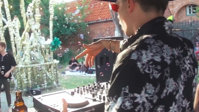A DJ wearing a floral shirt plays a live set at an outdoor garden party with floating bubbles.