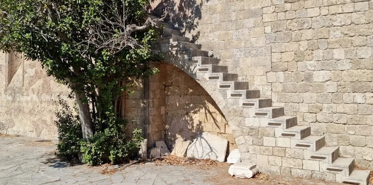 Ancient stone steps built into a medieval fortress wall next to a green tree in Rhodes, Greece.