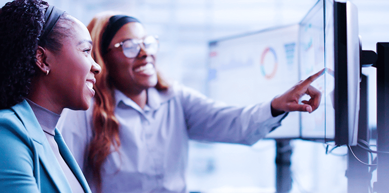 Two business professional women analyzing data charts and analytics on computer monitors.
