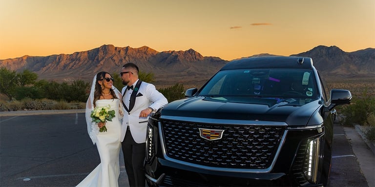 A bride and groom pose by a black Cadillac Escalade in the desert at sunset.