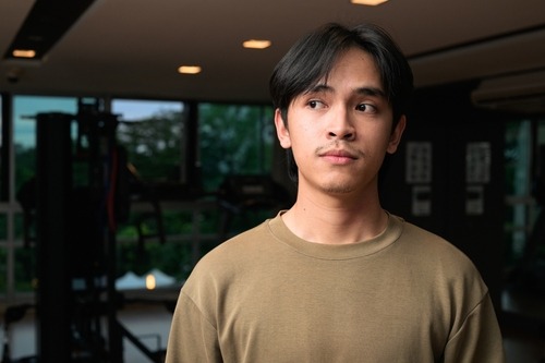A young man in a tan sweatshirt standing in a modern gym with exercise equipment in the background.