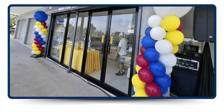 A festive entrance with colorful balloon arches beside glass doors, a table with a yellow cloth, and a person setting up