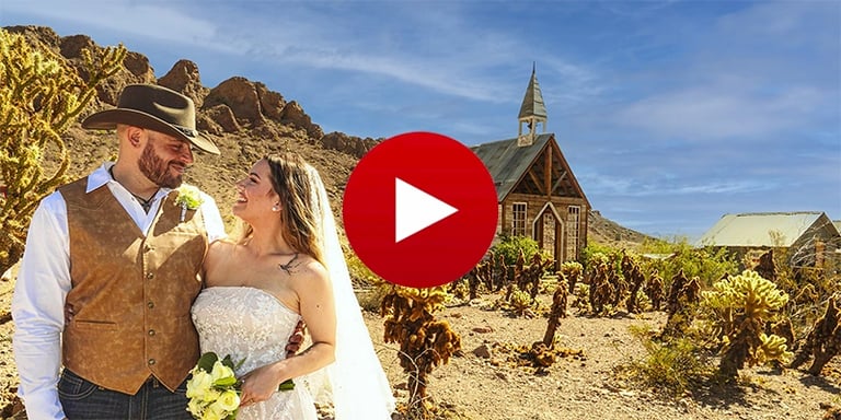 A smiling couple at a desert wedding in front of a rustic chapel and cactus plants.