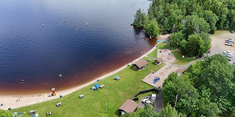 Aerial view of a public beach park with a grassy picnic area, playground, and people kayaking on the lake.