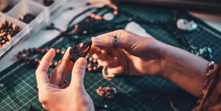 Artesana de joyería inspeccionando una piedra natural oscura en su mesa de trabajo con herramientas y cuentas de macramé.