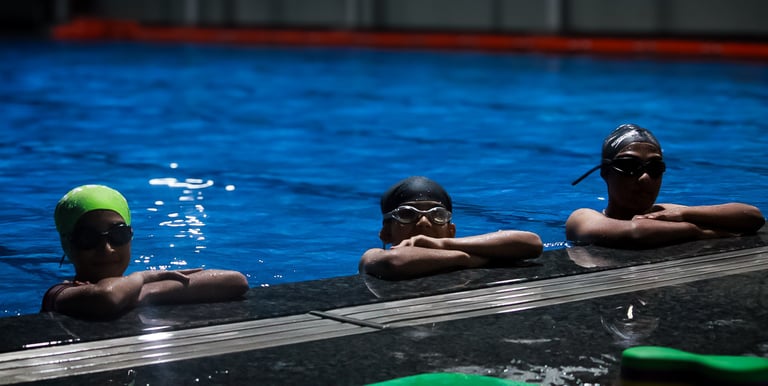 “Children wearing swim caps and goggles resting at the edge of a swimming pool.”