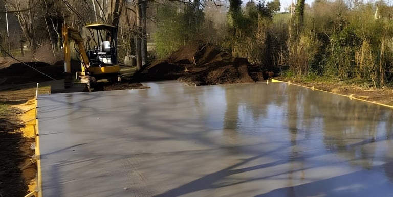 A freshly poured wet concrete slab foundation at a construction site with an excavator in the background.