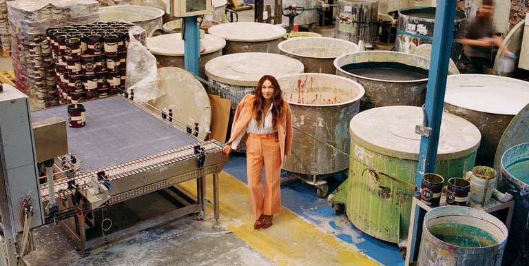 A woman in a coral suit standing in a professional paint factory with industrial mixing vats and conveyors.