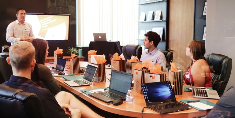 Colleagues attending a corporate business meeting in a modern office boardroom with laptops and a presenter.